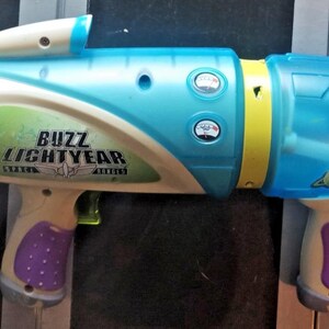 May include: A Buzz Lightyear toy water gun in shades of blue, white, yellow, and purple. The toy features the Buzz Lightyear logo and character image. It has a red dial and a trigger, designed for water play.