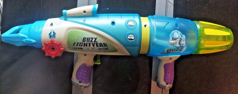 May include: A Buzz Lightyear toy water gun in shades of blue, white, yellow, and purple. The toy features the Buzz Lightyear logo and character image. It has a red dial and a trigger, designed for water play.