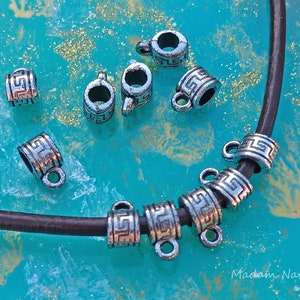 May include: Silver-colored metal jewelry findings with a Greek key design. The findings are cylindrical with a loop attached, and are shown on a dark brown leather cord against a turquoise background.