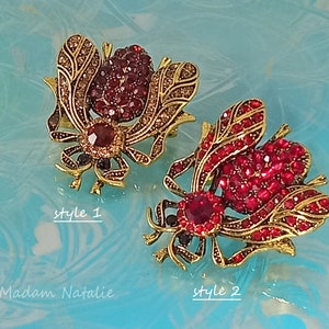May include: Two gold-toned bee brooches with red and clear rhinestones. The bee on the left has a brown center and the bee on the right has a red center. The bee on the left is labeled "style 1" and the bee on the right is labeled "style 2".