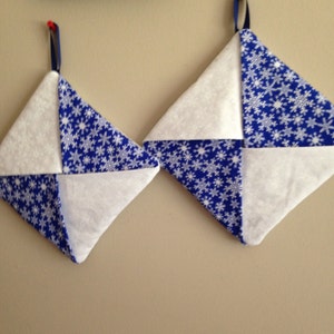 May include: Two blue and white pot holders with a snowflake pattern. The pot holders are hanging on a wall.
