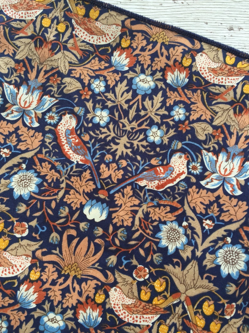 May include: Close-up of a fabric with a repeating pattern of birds, flowers, and foliage in shades of navy blue, orange, tan, and light blue. The design features stylized birds with red and white markings, surrounded by floral and leaf motifs.