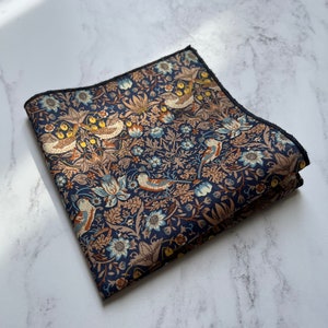 May include: Folded navy blue pocket square with a detailed floral and bird pattern in shades of tan, blue, and yellow. The edges are finished with a dark border. The fabric is likely cotton or a similar material.