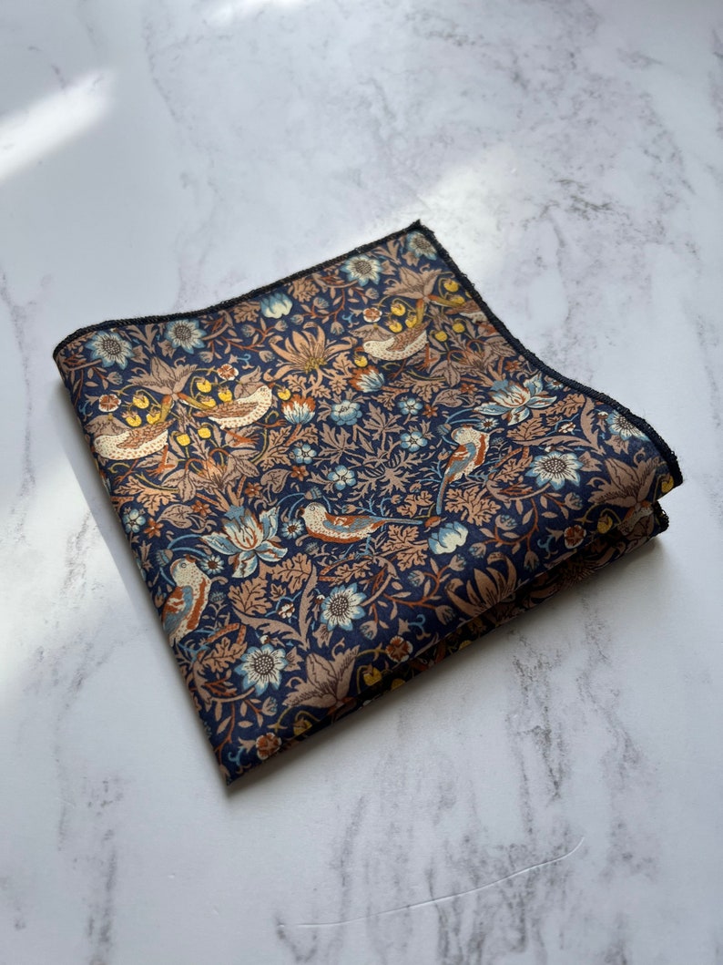May include: Folded navy blue pocket square with a detailed floral and bird pattern in shades of tan, blue, and yellow. The edges are finished with a dark border. The fabric is likely cotton or a similar material.