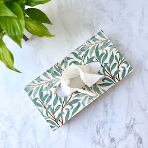 May include: A rectangular tissue box with a botanical design of blue-green leaves and brown branches on a cream background. White tissues emerge from the oval opening. A green plant is in the upper left corner.