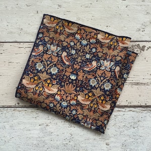 May include: A navy blue pocket square with a detailed floral and bird pattern. The design features birds, flowers, and foliage in shades of brown, orange, and blue. The edges are finished with a dark blue border.