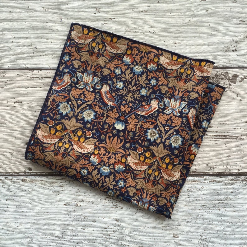 May include: A navy blue pocket square with a detailed floral and bird pattern. The design features birds, flowers, and foliage in shades of brown, orange, and blue. The edges are finished with a dark blue border.