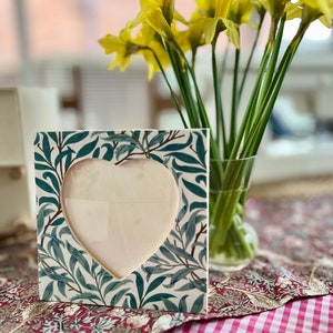 May include: A heart-shaped picture frame with a green and white floral pattern. The frame is sitting on a red and white checked tablecloth with a bouquet of yellow daffodils in a glass vase.