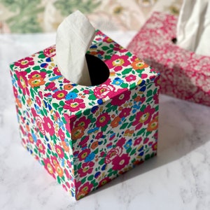 May include: A square tissue box with a floral pattern in shades of pink, blue, green, and orange. The box is made of cardboard and has a white tissue dispenser.