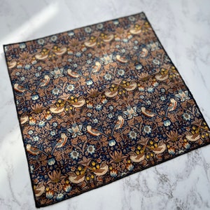 May include: Square navy blue handkerchief with a detailed floral and bird pattern. The design features stylized birds, flowers, and foliage in shades of blue, brown, and cream. The edges are finished with a black border.