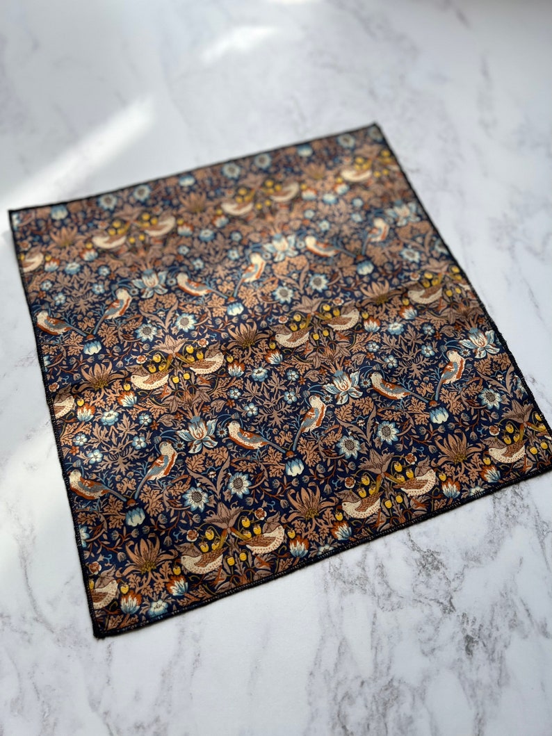 May include: Square navy blue handkerchief with a detailed floral and bird pattern. The design features stylized birds, flowers, and foliage in shades of blue, brown, and cream. The edges are finished with a black border.