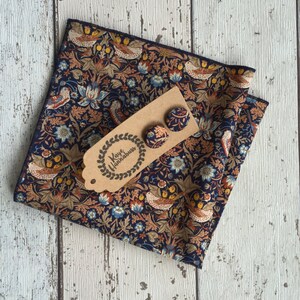 May include: A navy blue floral patterned pocket square and matching stud earrings. The fabric features a repeating design of birds, flowers, and foliage in shades of orange, blue, and brown. The earrings are attached to a brown tag with the text "Kaye Hannabus".