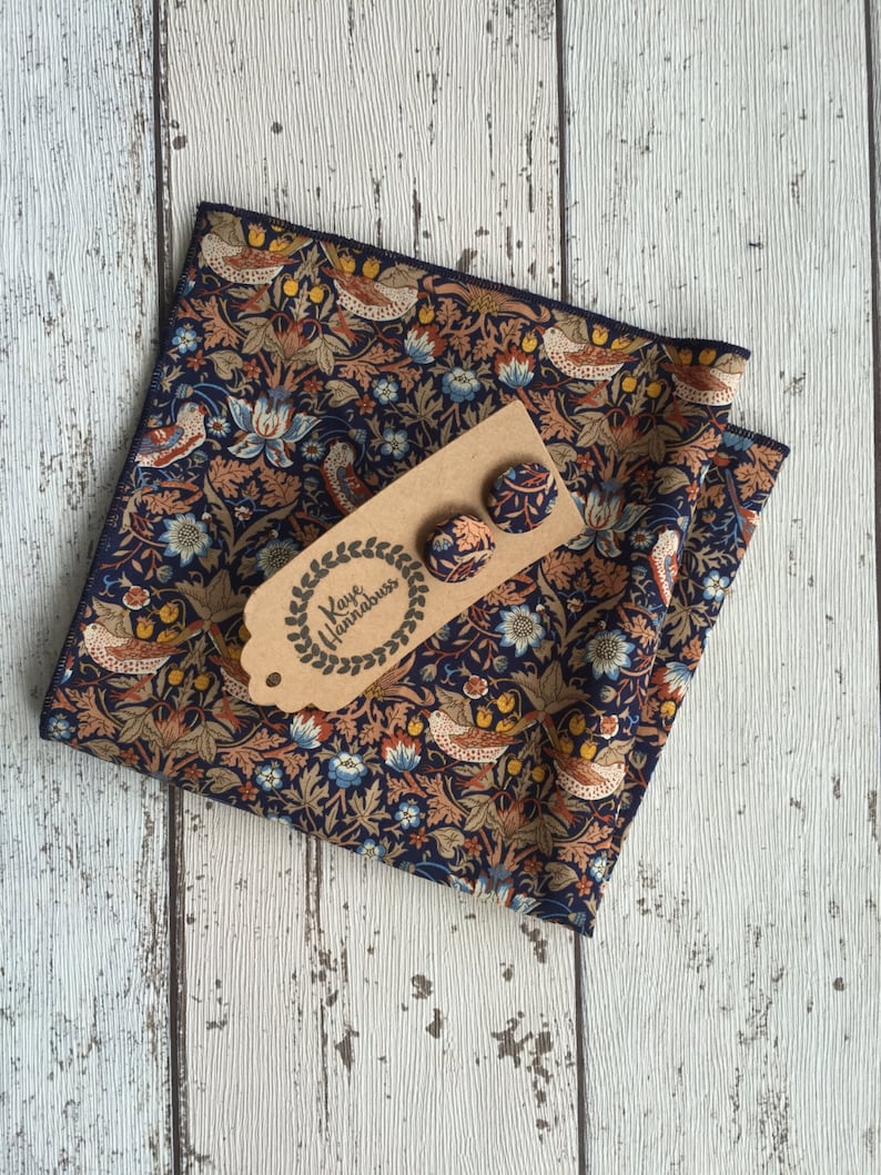 May include: A navy blue floral patterned pocket square and matching stud earrings. The fabric features a repeating design of birds, flowers, and foliage in shades of orange, blue, and brown. The earrings are attached to a brown tag with the text "Kaye Hannabus".