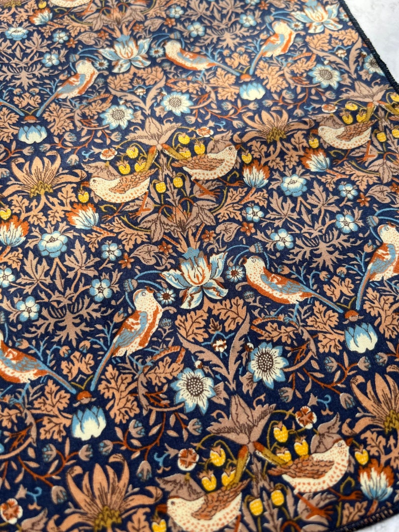 May include: A navy blue fabric with a repeating pattern of birds, flowers, and foliage in shades of brown, beige, and blue. The design features detailed illustrations of birds perched among stylized floral elements. The overall aesthetic is reminiscent of vintage textile designs.