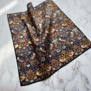 May include: A folded square cloth with a detailed floral and bird pattern in shades of navy blue, brown, and cream. The fabric has a black border and is likely a handkerchief or decorative cloth.