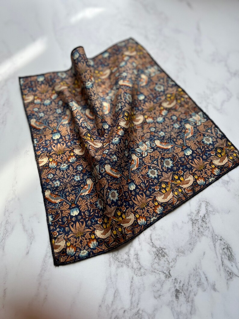 May include: A folded square cloth with a detailed floral and bird pattern in shades of navy blue, brown, and cream. The fabric has a black border and is likely a handkerchief or decorative cloth.