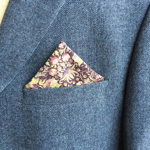 May include: A blue tweed blazer with a floral patterned pocket square. The pocket square features a dark background with colorful flowers and leaves. The blazer has a classic design.