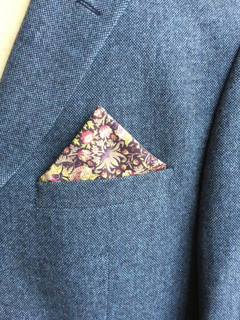 May include: A blue tweed blazer with a floral patterned pocket square. The pocket square features a dark background with colorful flowers and leaves. The blazer has a classic design.