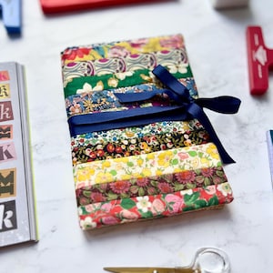 May include: A collection of patterned fabric strips in a variety of floral designs, tied with a navy blue ribbon. The fabrics display a mix of colours including red, yellow, green, and blue. Gold scissors and stationery items are in the background.