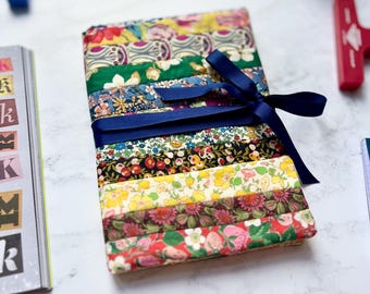 May include: A stack of fabric strips in various floral patterns, bound with a navy blue ribbon. The fabrics feature a mix of colors including red, yellow, green, and blue. A pair of gold scissors and other stationery items are in the background.