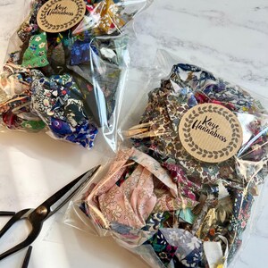 May include: Two clear bags of fabric scraps, each with a "Kaye Hannabuss" sticker. The fabric pieces are in various colours and patterns, including floral and geometric designs. A pair of black scissors is placed nearby.