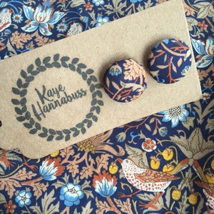 May include: Two round stud earrings with a floral pattern in navy blue, peach, and light blue, resting on a kraft paper tag with the text "Kaye Hannabuss". The earrings are displayed on a matching fabric with a similar floral design.