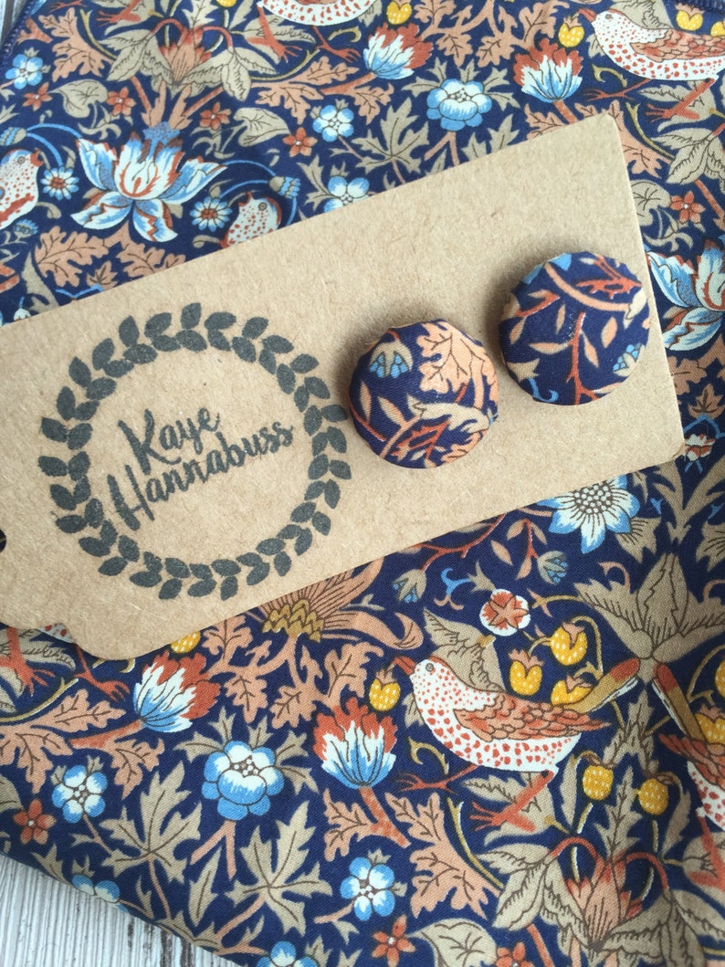 May include: Two round stud earrings with a floral pattern in navy blue, peach, and light blue, resting on a kraft paper tag with the text "Kaye Hannabuss". The earrings are displayed on a matching fabric with a similar floral design.