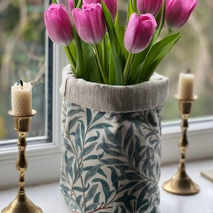 May include: A bouquet of pink tulips in a fabric planter with a willow pattern. The planter is sitting on a windowsill with two gold candle holders.