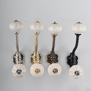 May include: Four wall hooks featuring white ceramic knobs with a beige petal pattern. Each hook has a twisted metal stem and a floral-shaped mounting plate. The metal finishes include silver, gold, bronze, and black, each with a matching center screw.
