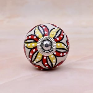 May include: A round ceramic cabinet knob featuring a hand-painted floral pattern with alternating red and yellow petals outlined in black. The design includes small white raised dots along the petals and a central silver-toned metal hardware piece.