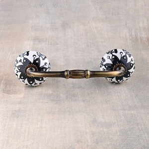 Könnte beinhalten: Schwarz-weißer Keramik-Schubladengriff mit dekorativem Blumendesign. Der Griff hat eine gebogene Metallstange, die die beiden Keramikknöpfe verbindet.