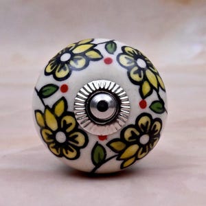 May include: A round ceramic cabinet knob with a white base featuring hand-painted yellow flowers with black outlines, green leaves, and small red dots. The centre of the knob has a silver-coloured metal circular plate with a ridged edge and a screw hole.