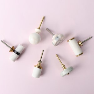 Op de afbeelding: Zes verschillende marmeren kastknoppen met gouden accenten. De knoppen zijn in verschillende vormen, waaronder rond, vierkant en cilindrisch.