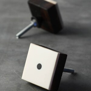 Könnte beinhalten: Zwei quadratische Schrankknöpfe mit schwarzem Holzrahmen und weißem Zentrum. Ein Knopf hat einen schwarzen Punkt in der Mitte.