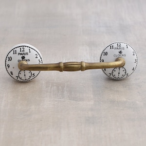 以下が含まれることがあります： 真鍮製の引き出しの取っ手に、白い磁器製の時計の文字盤のノブが2つ付いています。時計の文字盤には黒い針と黒いローマ数字があります。時計の文字盤には黒い文字で「Paris Clocks」と書いてあります。