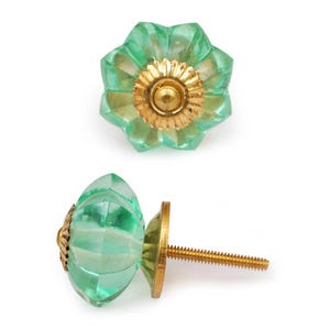 May include: Two decorative cabinet knobs. The knobs are a light green colour with a faceted, flower-like design. The centre of the knobs and the base are gold-coloured. One knob is shown from the front, and the other is shown from the side.