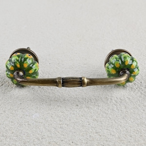 May include: A green and yellow ceramic drawer pull with a brass bar connecting the two knobs.