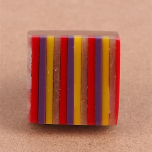 Könnte beinhalten: Ein quadratischer Ring mit einem farbenfrohen Streifendesign. Der Ring hat vertikale Streifen in Rot, Gelb und Lila, eingebettet in transparentem Harz. Ein auffälliges Schmuckstück.