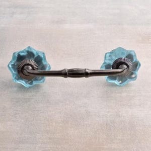 Puede incluir: Un par de pomos de armario de vidrio azul claro con una barra de conexión de metal plateado. Los pomos tienen un diseño de estrella.