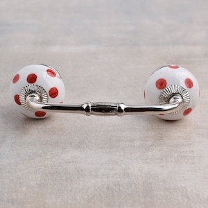 May include: White ceramic drawer pull with red polka dots. The pull has a silver metal bar connecting the two knobs.