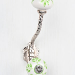 May include: A silver metal hook with a twisted stem and two white ceramic knobs with green leaf designs.