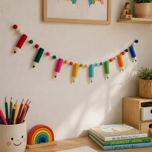 May include: A felt garland featuring miniature pencil shapes in red, dark green, pink, yellow, light blue, lime green, and orange, interspersed with small multicolored felt balls, hanging horizontally against a plain white wall.