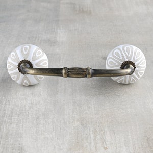 May include: White ceramic drawer pull with a decorative pattern and a brass bar connecting the two knobs.