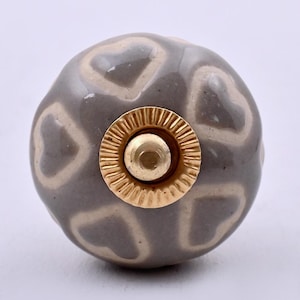 May include: A round ceramic cabinet knob featuring a glossy grey finish with a repeating pattern of light beige heart shapes. The center of the knob is accented with a circular, ridged gold-toned metal hardware piece with a smooth, rounded top.
