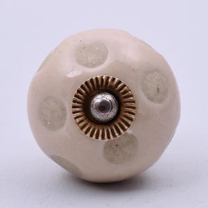 May include: A round, light beige ceramic cabinet knob featuring a pattern of pale, circular spots. The centre of the knob is accented with a metallic, fluted gold-toned washer surrounding a small, silver-coloured circular screw head attachment.