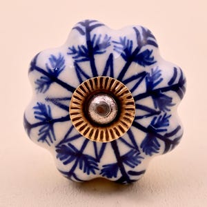 May include: A flower-shaped ceramic cabinet knob featuring a white base with a dark blue painted botanical pattern. The center of the knob has a circular, ridged gold-toned metal plate with a small silver-toned circular accent in the middle.