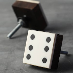 May include: Two square cabinet knobs with a wooden frame and a white face with black dots. The knobs are shaped like dice with different numbers of dots on each face.