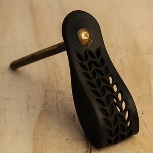 May include: Black leather cabinet pull with a leaf design and a brass screw. The pull has a cutout design and is attached to a metal screw.