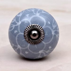 Op de afbeelding: Een ronde, lichtblauwe keramische meubelknop met een wit, met de hand geschilderd bloemmotief. Het midden van de knop is voorzien van een cirkelvormig, geribbeld zilverkleurig metalen beslag met een gladde, reflecterende koepel.