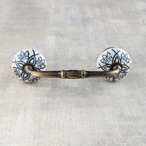 Può includere: Una maniglia per cassetto in ottone con due pomelli in ceramica bianca. I pomelli hanno un motivo floreale blu.